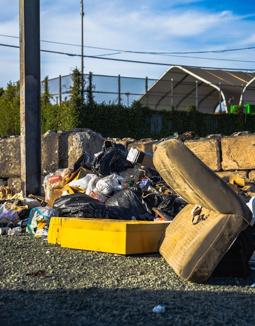 A pile of mixed household rubbish and waste materials accumulated on an outdoor gravel surface near a stone wall, featuring black plastic rubbish bags, a yellow plastic storage box, and an old, dirty car tire leaning against the debris. The background includes a metal fence topped with greenery, with a tennis court or sports facility partially visible behind it, along with utility poles and overhead power lines against a partly cloudy sky. The scene suggests a private or alternative rubbish collection setup, with visible waste that may be slated for disposal or removal by a professional waste management company such as Rubbish Removal Lewisham, highlighting the need for efficient rubbish clearance services in the area.