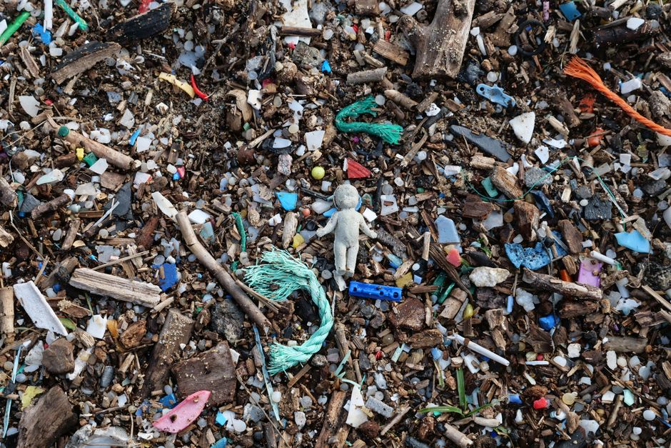 A close-up view of a section of ground covered with a mixture of small debris and waste materials, including fragments of plastic, bits of wood, and dirt. Dominant objects include a small, worn, light gray plastic figurine resembling a human shape, positioned centrally among the scattered debris. Surrounding the figurine are various pieces of plastic in different colors such as blue, pink, and green, along with a frayed piece of turquoise string lying nearby. Piled among the debris are small twigs, soil, and gravel, creating a textured and uneven surface. The scene appears to be outdoors, possibly on a dirt or gravel surface, with natural lighting emphasizing the mixture of organic and synthetic waste. The composition suggests an area that has undergone some form of rubbish clearing, highlighting the types of waste encountered in private or independent rubbish removal services like those offered by Rubbish Removal Lewisham.
