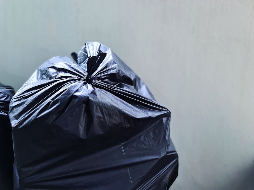 A pile of mixed household rubbish and waste materials accumulated on an outdoor gravel surface near a stone wall, featuring black plastic rubbish bags, a yellow plastic storage box, and an old, dirty car tire leaning against the debris. The background includes a metal fence topped with greenery, with a tennis court or sports facility partially visible behind it, along with utility poles and overhead power lines against a partly cloudy sky. The scene suggests a private or alternative rubbish collection setup, with visible waste that may be slated for disposal or removal by a professional waste management company such as Rubbish Removal Lewisham, highlighting the need for efficient rubbish clearance services in the area.