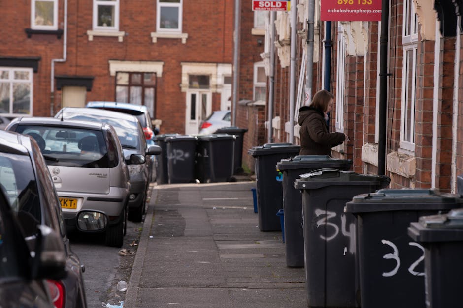 A suburban residential street scene showing a row of dark grey wheelie bins positioned along the pavement outside brick terraced houses. The bins are arranged in a line, some with open lids exposing waste inside, and are marked with white spray-painted numbers. In the background, a woman dressed in a brown coat is seen opening one of the bins, possibly for rubbish disposal or collection. The street features parked cars on the left side, including a silver hatchback closest to the camera, and extends into more residential buildings with red brick facades and white-framed windows. The environment appears well-lit with natural daylight, and the scene is typical of areas where private rubbish collection services, such as those offered by Rubbish Removal Lewisham, might operate as an alternative to council waste collection, supporting on-site or independent rubbish disposal.