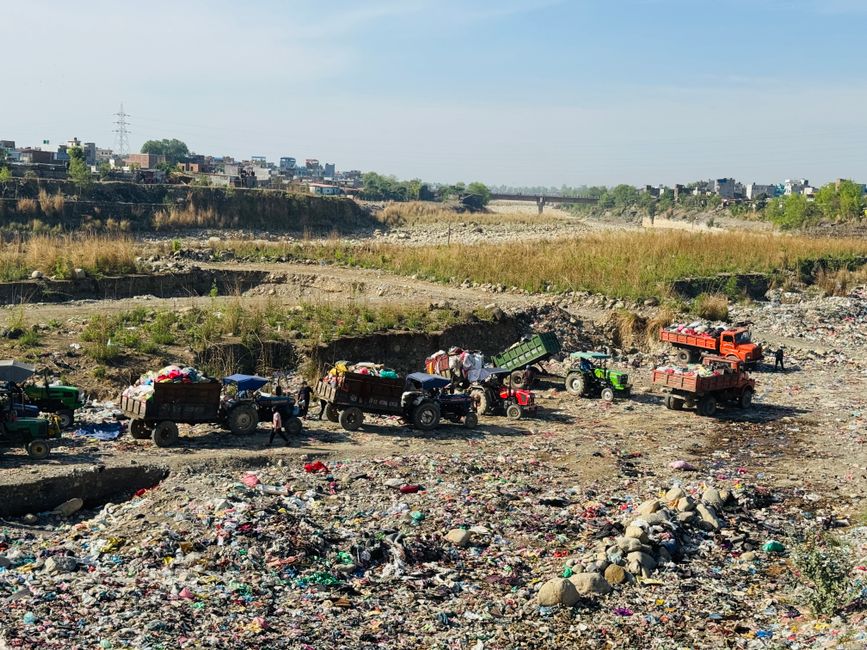 A large open area of land filled with scattered waste and rubbish, including plastic bags, bottles, and miscellaneous debris, with several small tractors and trucks parked across the site. The machinery appears to be used for moving and collecting waste materials within the area. The ground is uneven and rocky, with some patches of dry grass and exposed soil. In the background, there is a distant view of a residential neighborhood with low-rise buildings and trees, under a clear sky with soft sunlight illuminating the scene. The overall environment suggests a waste disposal or clearing site, potentially operated by an independent waste management service such as Rubbish Removal Lewisham, or a similar private collection operation engaged in on-site clearance of rubbish. The scene underscores the importance of effective waste handling methods outside standard municipal rubbish collection, reflecting the context of alternative rubbish removal solutions.