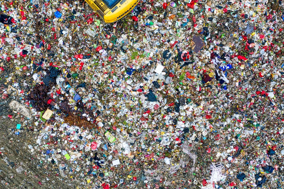 An expansive aerial view of a large land area covered with a dense layer of mixed waste, including plastic bottles, wrappers, paper, fabric, and other household refuse. The surface appears muddy and uneven, with areas of accumulated debris forming small mounds and patches of scattered waste. In the top right corner, a yellow refuse collection truck is partially visible, indicating ongoing rubbish removal activity, while the surrounding environment suggests an outdoor site, possibly designated for waste disposal or temporary storage. The waste materials are varied in color, predominantly white, red, blue, and black, with some organic matter and vegetation interspersed among the garbage. The scene underscores the scale of unmanaged or overflowing refuse, aligning with issues often addressed through private rubbish removal services such as those provided by Rubbish Removal Lewisham, especially in contexts involving atypical or large-scale waste clearance outside standard municipal collection systems.