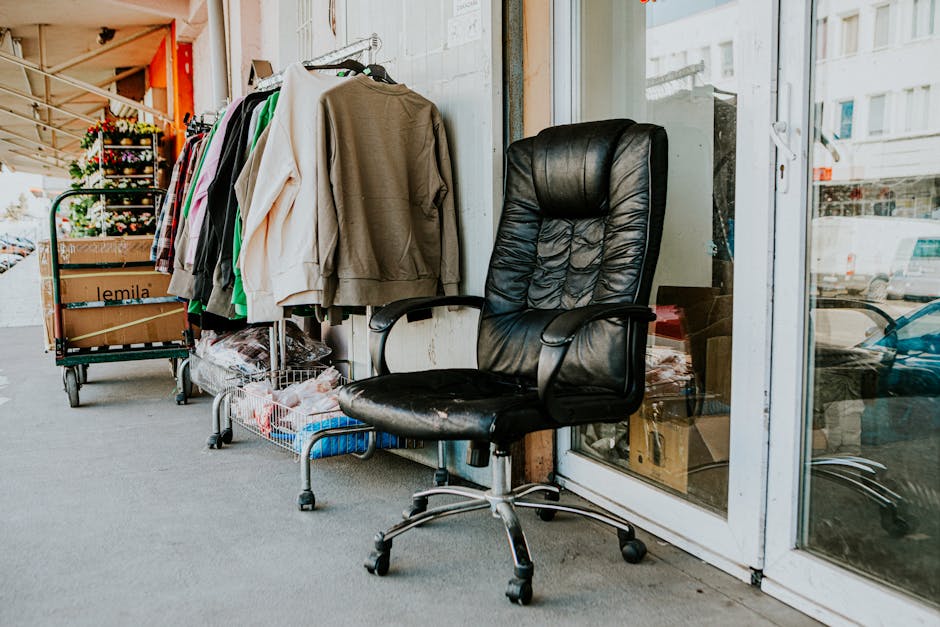 The image shows an outdoor area adjacent to a building, with a black leather office chair placed on a concrete surface in the foreground. The chair has a high backrest, padded seat, and armrests, and appears slightly worn with some creases in the leather. Behind the chair, there is a large window or glass door reflecting nearby parked vehicles and urban surroundings. To the left of the chair, a metal clothing rack is positioned against the building's wall, holding a variety of garments including jackets, sweatshirts, and shirts in different colours and fabrics, some hanging on hangers and others draped over the rack. Below the clothing, a small cart or trolley with wheels contains miscellaneous items, possibly textiles or packaging materials. The setting suggests a commercial or retail environment, possibly part of a shop or storage area, where discarded clothing and miscellaneous waste items are temporarily placed for collection or removal, reflecting the kinds of materials handled by a waste management service such as Rubbish Removal Lewisham dealing with private or independent rubbish collection. The natural daylight and urban background add to the everyday scene of waste or item clearance outside a commercial building.