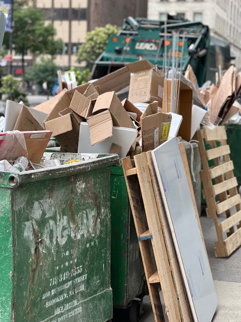 A large green waste containment bin filled with various types of discarded rubbish, including cardboard boxes, flattened cardboard packaging, some white insulative foam, and wooden pallets leaning against the side of the bin. The cardboard boxes are of different sizes and some are open or partially crushed, revealing internal flaps and packaging layers. The wooden pallets are light-colored, with visible slats and nails, and positioned vertically beside the bin on a paved urban street. In the background, a recycling truck with a dark green body, hydraulic arms, and orange safety lights is partially visible, parked on the street amidst a cityscape with trees and multi-storey buildings. The scene appears to be in daylight with natural lighting, typical of an urban waste collection area suitable for private rubbish removal or alternative waste handling services like those offered by Rubbish Removal Lewisham, illustrating the process of clearing bulky waste or large-volume rubbish in a city environment.