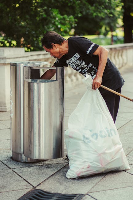 A middle-aged man with short dark hair is seen disposing of rubbish into a stainless steel public trash bin on a paved outdoor walkway. He is bending slightly forward, wearing a black T-shirt with white and blue graphic text on the front, and holding a white plastic bag filled with waste in his left hand, while his right hand is reaching into the open-top container. The ground is tiled with grey paving slabs, and in the background, there is a low stone balustrade with lush green trees behind it, suggesting a park or public space setting. The scene is illuminated by natural daylight, highlighting the reflective surface of the trash bin and the man's focused posture, consistent with practices of proper waste disposal often associated with community cleanliness and waste management efforts, as provided by Rubbish Removal Lewisham.