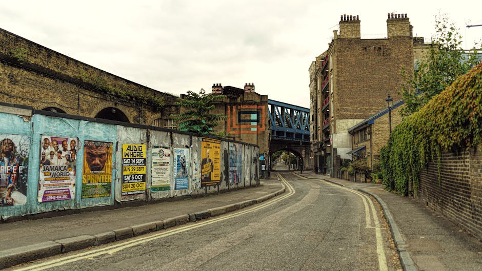 A narrow urban street scene featuring a slightly curved asphalt road with double yellow lines along the right edge, bordered by a brick wall covered in green ivy on the right side and a weathered concrete wall on the left, which is decorated with numerous colorful posters and advertisements. In the background, a blue metal bridge spans above the street, supported by brick structures and stone archways, with a mix of multi-story brick buildings featuring chimneys and balconies on either side. The sky above is overcast, casting diffuse light over the scene, creating a muted, neutral tone. The environment appears to be part of a city area, with typical street-level clutter that might be associated with local community notices or promotional events, aligning with the context of alternative waste handling and independent collection efforts often seen in urban rubbish removal settings. This scene visually supports the idea of private on-site clearance or disposal in an inner-city environment managed by services like Rubbish Removal Lewisham.