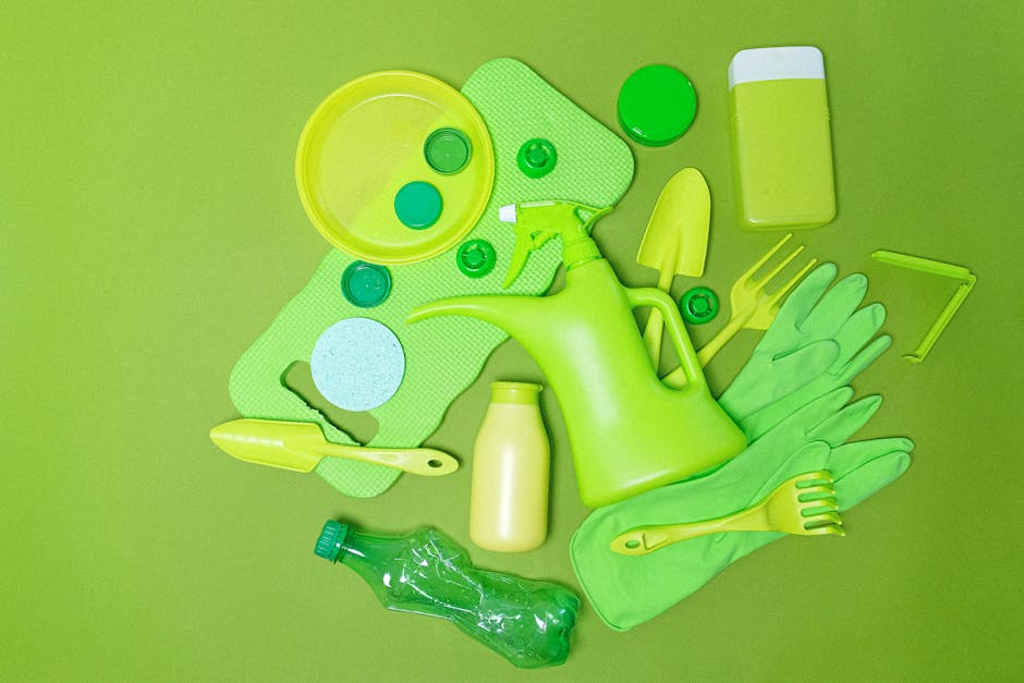 A collection of green cleaning and gardening tools is laid out on a plain green surface, including a watering can, rubber gloves, a spray bottle, a small trowel, a plastic bottle, a scrub brush, a sponge, a soap or detergent container, and a plastic scraper. The tools are made of plastic or rubber, with textures ranging from smooth to slightly textured surfaces, predominantly in various shades of green with some yellow accents. The watering can is centrally positioned, with the gloves placed to its right, and the spray bottle and small trowel near the bottom left and right sides respectively. The sponge, soap container, and scraper are distributed across the scene, creating a neatly arranged display of domestic cleaning and gardening implements. The even lighting highlights the colors and textures, emphasizing their material qualities, and the overall composition subtly reflects the type of items typically prepared or sorted for waste collection or disposal services such as those offered by Rubbish Removal Lewisham, supporting indoor or outdoor rubbish management activities.