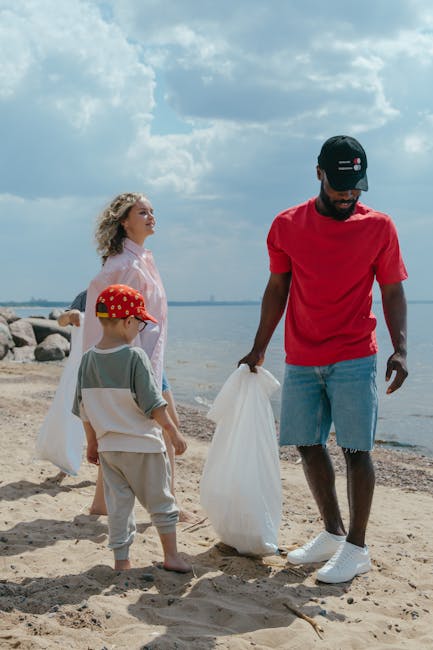 A group of three people, including a man and two children, are participating in a beach clean-up activity along the shoreline. The man, positioned to the right, is wearing a red T-shirt, light blue denim shorts, and a black cap, and is holding a white waste bag, appearing to gather litter from the sand. The two children, standing to his left, include a young boy wearing a white and gray outfit with a red cap decorated with yellow dots, and a girl in a pink patterned shirt with curly blonde hair, watching attentively. The sandy beach extends toward a rocky shoreline in the background, with calm water and a partly cloudy sky overhead. The scene indicates a community effort focused on environmental cleanup, aligned with the principles of rubbish removal and private waste handling services such as those offered by Rubbish Removal Lewisham, emphasizing on-site clearance and alternative waste collection methods.
