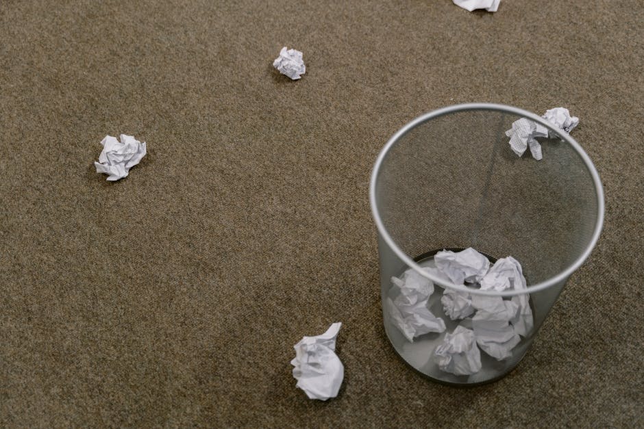 A clear cylindrical waste bin made of semi-transparent plastic is positioned on a brown carpeted floor, approximately in the lower right quadrant of the image. The bin contains several crumpled sheets of white paper, with some protruding above the rim. Additional crumpled paper pieces are scattered across the carpet, lying randomly nearby the bin. The carpet has a textured appearance with a mix of darker and lighter brown fibers. The lighting appears even and natural, without strong shadows, creating a neutral and professional atmosphere typical of an indoor setting. The scene reflects an environment focused on waste disposal or office rubbish collection, which aligns with the context of private waste handling services provided by Rubbish Removal Lewisham, and visually suggests the task of collecting and managing miscellaneous paper waste in a professional or domestic space.