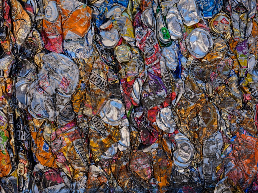 A large pile of crushed aluminium cans and beverage containers, primarily silver in color, with some painted in various bright colors including red, green, and orange. The cans are densely crumpled, overlapping each other, and exhibit different levels of sheen, reflecting light with a metallic finish. The pile appears to be on a flat surface, possibly outdoors or in a waste collection area, with no visible background or other objects nearby. The cans display torn labels and branding, indicating they are discarded drink containers. This scene illustrates waste comprised of recyclable aluminium in preparation for collection or recycling processes, relevant to private waste handling or rubbish removal services such as those provided by Rubbish Removal Lewisham.