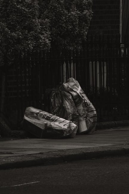 The image depicts three large black plastic garbage bags, placed on the pavement next to a black metal fence with vertical bars, which runs horizontally across the background. The bags are filled with waste, appearing somewhat lumpy and crinkled, with visible shiny surfaces reflecting minimal light. Behind the fence, parts of the dense foliage of a bush or small tree are visible, adding a natural backdrop to the scene. The setting appears to be outdoors, possibly on a street or alleyway, with a dark, overcast atmosphere illuminated by diffuse natural light. The bags are positioned close to the curb, suggesting an informal rubbish disposal collection, consistent with private waste handling services like those offered by Rubbish Removal Lewisham, to facilitate efficient on-site clearance or alternative waste management without reliance on public waste collection.