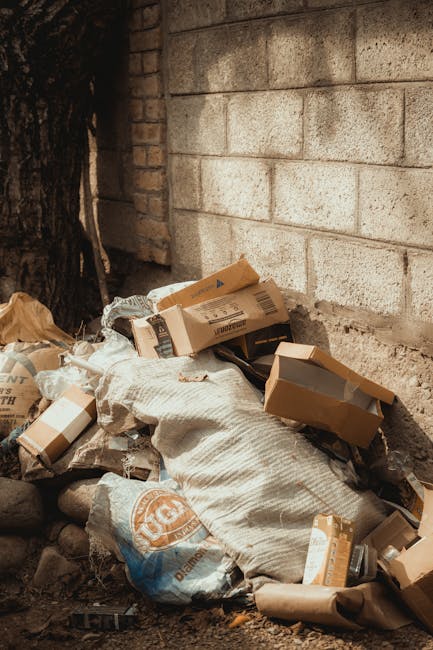 The image depicts three large black plastic garbage bags, placed on the pavement next to a black metal fence with vertical bars, which runs horizontally across the background. The bags are filled with waste, appearing somewhat lumpy and crinkled, with visible shiny surfaces reflecting minimal light. Behind the fence, parts of the dense foliage of a bush or small tree are visible, adding a natural backdrop to the scene. The setting appears to be outdoors, possibly on a street or alleyway, with a dark, overcast atmosphere illuminated by diffuse natural light. The bags are positioned close to the curb, suggesting an informal rubbish disposal collection, consistent with private waste handling services like those offered by Rubbish Removal Lewisham, to facilitate efficient on-site clearance or alternative waste management without reliance on public waste collection.