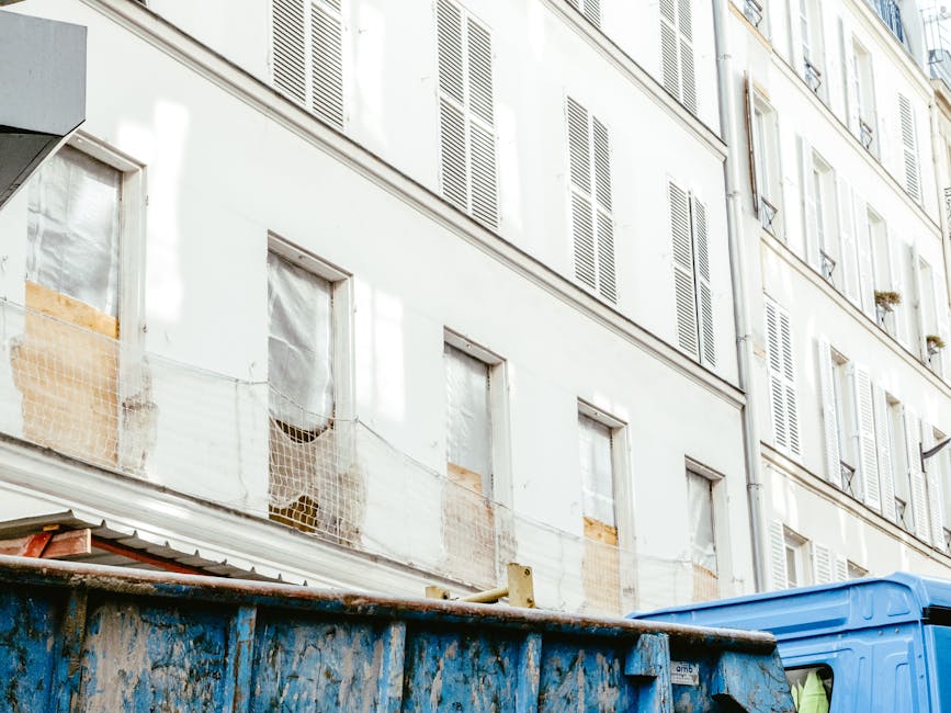The image shows a multi-storey residential building with a white facade, multiple windows with white shutters, and some windows covered with protective plastic and netting, indicating ongoing renovation or refurbishment. In the foreground, a large blue skip container is positioned on a paved surface, filled with various discarded materials including wooden planks and construction debris. The skip exhibits signs of weathering with rust and dirt on its surface, and its open top reveals informal waste storage. Adjacent to the skip, part of a white delivery or waste collection truck is visible, suggesting an active rubbish collection or clearance process. The setting appears to be an urban environment, possibly in a residential or apartment district, where private waste disposal via skips and local collection is common. The overall scene emphasizes the use of on-site clearance methods as an alternative to traditional household rubbish collection, aligning with independent waste handling practices supported by companies such as Rubbish Removal Lewisham, while highlighting the typical materials and equipment involved in such services.