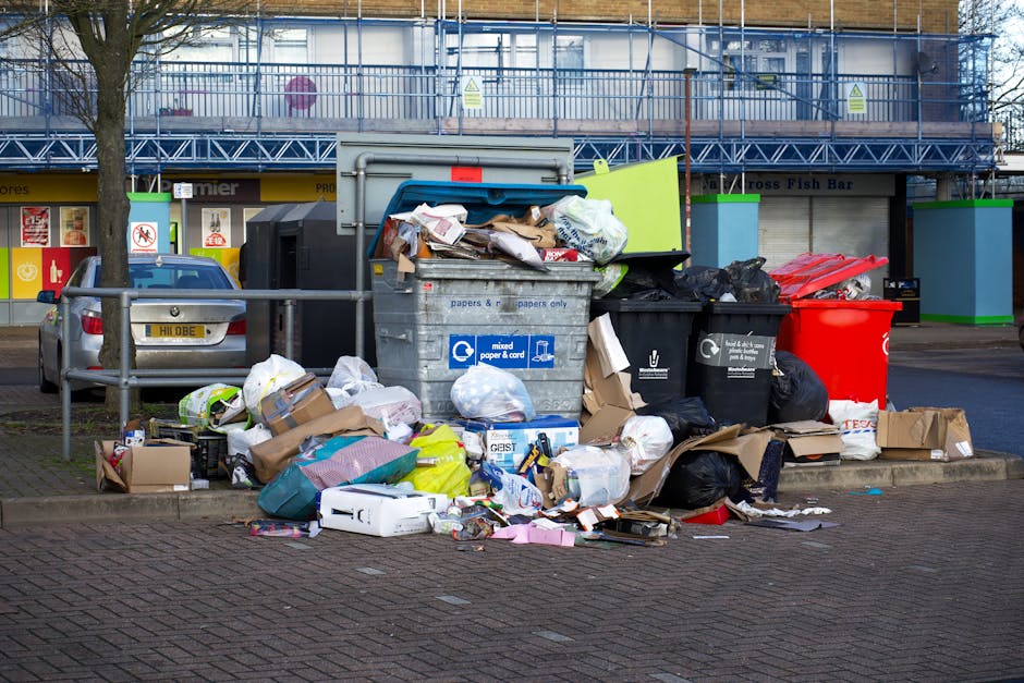 A large pile of mixed waste and rubbish overflowing from and surrounding a grey, metal recycling bin labeled for paper and cardboard, situated on a paved urban street. The debris includes crumpled cardboard boxes, plastic bags, paper parcels, and various household packaging materials, some spilling onto the surrounding area. To the left of the bin, there is a black trash bag and a red waste container. Nearby, a small silver car is parked behind a metal railing that separates the waste area from the sidewalk. In the background, a building with a blue metal balcony or scaffolding, storefront signage, and a few trees are visible under overcast lighting. The scene reflects instances where independent waste disposal or rubbish collection services may be used instead of municipal rubbish removal, aligning with private skip hire or on-site clearance practices often seen in waste management services such as Rubbish Removal Lewisham.
