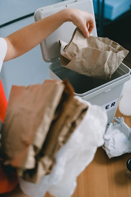 A person is placing a crumpled brown paper bag into a white rubbish bin with a black interior, situated on a wooden surface. The person's hand is visible, holding the bag as they fold or insert it into the bin, which appears to be designated for waste collection. In the foreground, a bundle of additional crumpled paper bags is being held, partially obscuring the view of the bin. The background includes part of a light blue wall, with the scene illuminated by natural or soft artificial lighting, emphasizing the textures of the paper bags and the smooth surface of the bin. This setup suggests the process of disposing of household waste, fitting within the context of independent rubbish handling services provided by Rubbish Removal Lewisham, and exemplifies typical waste segregation before collection within domestic settings.