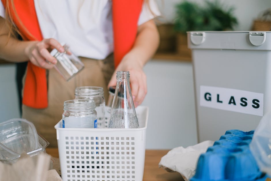 A person wearing a white shirt and red cardigan is seen from the waist up, handling a glass jar with their right hand while placing it into a white plastic basket that contains several other empty glass bottles and jars. The basket is positioned on a wooden surface, with a large grey container labeled 'GLASS' to the right, which is partially open with its lid resting on top. The background features indoor plants and wooden shelving, suggesting a household setting focused on waste sorting. The lighting is natural, highlighting the clear, smooth textures of the glass objects and contrasting with the matte finish of the plastic containers, emphasizing the importance of proper glass recycling and waste separation, a key aspect of independent waste handling and rubbish removal services such as those provided by Rubbish Removal Lewisham.